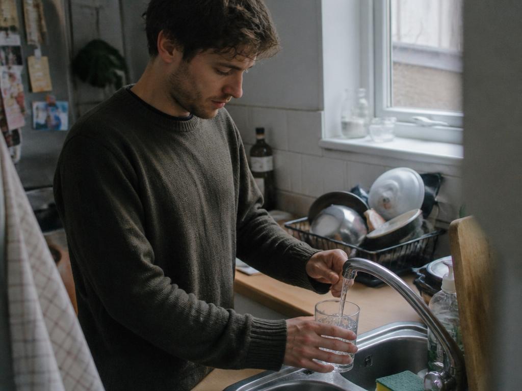 A man fills a plain pint glass with tap water at a kitchen sink, watching the running stream in cool overcast daylight.