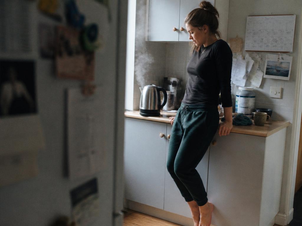 A woman leans against a kitchen counter doing a quick stretch while waiting for the kettle to boil in cool morning light.