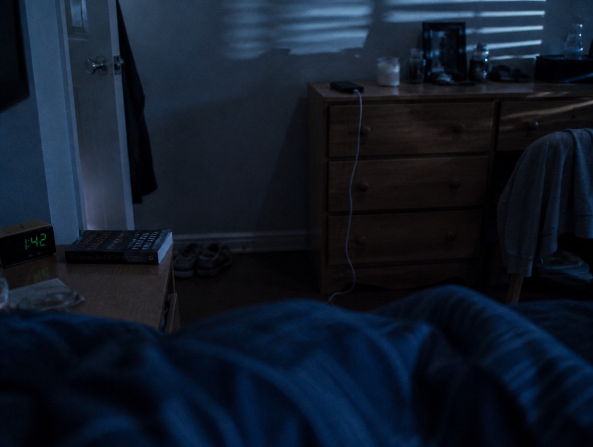 A dim bedroom at night lit by cool blue moonlight through blinds, showing a phone charging face-down on a dresser across the room while a digital clock and book sit on the nearby nightstand.